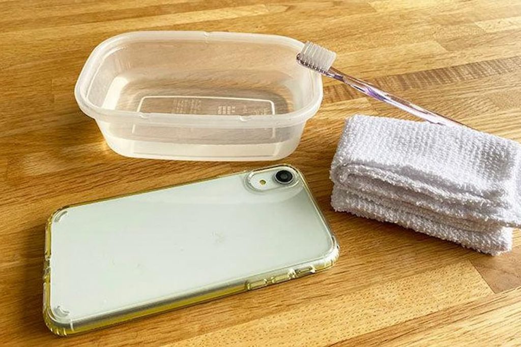 Essential tools for cleaning a phone case, including a bowl of water, a toothbrush, and a microfiber cloth on a wooden surface.